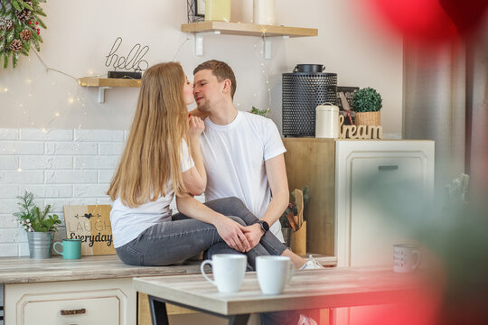 Couple in love kissing each other while sitting in kitchen at home in morning and drinking coffee.