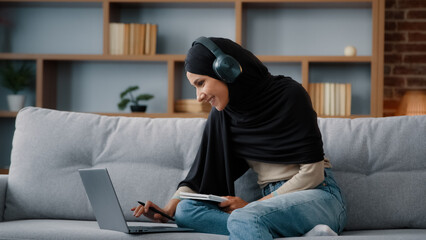 Islamic Arabian student girl Muslim woman wearing black hijab busy lady in headphones with laptop watching webinar lesson online class training write notes in notebook do homework e-learning at home