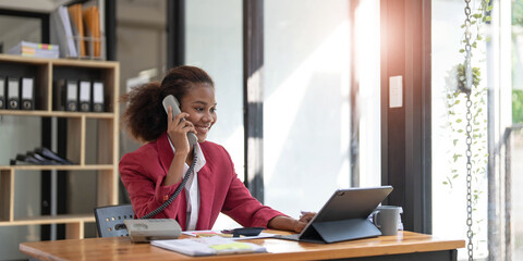 Happy african-american business woman talking on mobile at office. Young female manager consulting on phone, using laptop at working place, copy space
