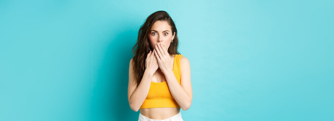 Portrait of attractive modern girl looks shocked after hear gossips, cover mouth with hands and gasping, staring with amazement at camera, standing over blue background - obrazy, fototapety, plakaty