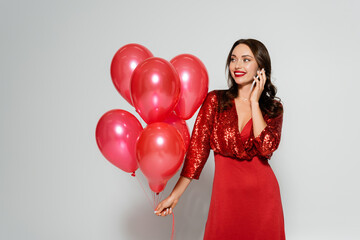 Cheerful woman in stylish red dress talking on cellphone and holding balloons on grey background
