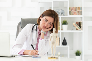 Portrait photo of young and beautiful thinking female doctor in white medical gown sitting and working on laptop in the office of the modern clinic. Online consultation and distant cure concept