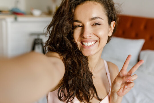 POV of happy Caucasian young brunette woman posing to smartphone camera taking selfie photo and smiling. Joyful positive beautiful lady takes picture of herself at home