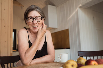Happy senior 60s woman wearing glasses sitting at the kitchen table