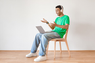 Excited arab man in wireless headphones making video call via laptop while sitting on chair over light wall, copy space