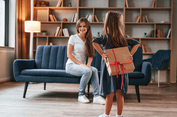 Girl holding gift box behind her. Happy holidays. Young mother with her daughter is together at apartment