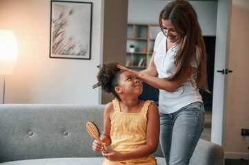 Mother is combs hair. Young woman with her daughter is together at apartment