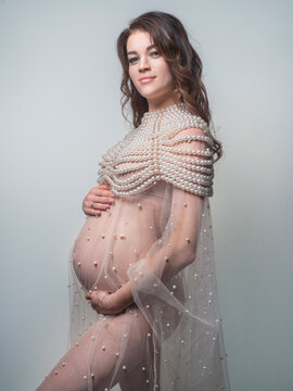 A tender photo of a young pregnant woman in a transparent dress decorated with pearls and a chic pearl necklace. Medium shot portrait on a gray background.