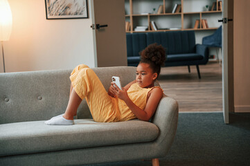 Surfing the internet by using smartphone. Cute black girl in casual clothes is at home at daytime