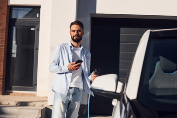 With smartphone. Young stylish man is with electric car at daytime