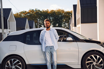 Standing and leaning on the vehicle. Posing for the camera. Young stylish man is with electric car...