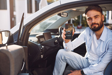 Sitting inside the vehicle, monitoring charging process by using smartphone. Young stylish man is with electric car