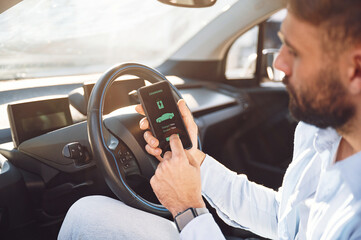 Monitoring charging procces by using smartphone. Young stylish man is with electric car at daytime