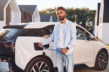 Plugging in charging cable. Young stylish man is with electric car at daytime