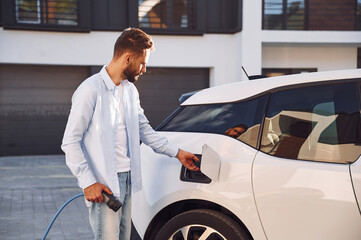 Plugging in charging cable. Young stylish man is with electric car at daytime