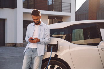 Charging the vehicle near the house. Holding smartphone. Young stylish man is with electric car at...