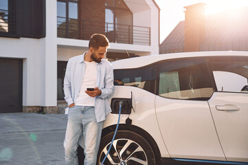 Charging the vehicle near the house. Holding smartphone. Young stylish man is with electric car at daytime - Powered by Adobe