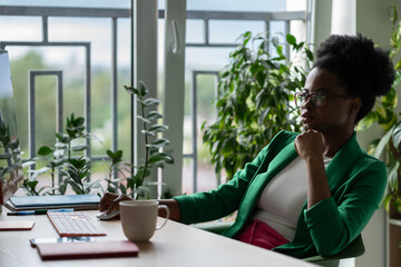Self-sufficient African American business woman sits at work desk with coffee cup and computer. Stylish girl manager works in office with green plants in confident pose checking or financial report