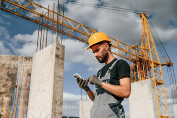 With phone in hands. Standing against crane. Man in uniform is working on the construction site