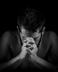 closeup of young shirtless man with beard praying with hands folded against black background...
