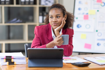 Smiling young African american female Finance Entrepreneur sitting in workplace office hoding coffee cup and working on a laptop, investment financial document report finance analysis concept.