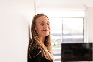 Close up shot of a smiling blonde woman with long hair wearing black blouse leaning on a white wall