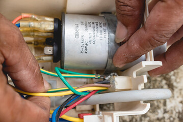 An electrician examines the wiring connected to CBB65A-1 capacitor inside a window type air conditioner control panel outside.