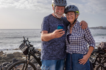 Smiling senior caucasian couple standing on the pebble beach close to their electric bicycles using...
