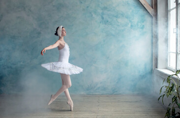Fototapeta na wymiar a professional ballerina in a white tutu is dancing in a room with blue wall and smoke