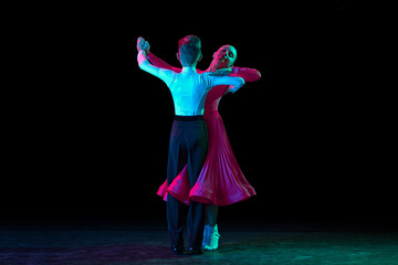 Waltz. Full-length portrait of beautiful little boy and girl dancing ballroom dance isolated over dark background in neon light. Concept of art, beauty, grace, action, emotions.