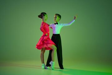 Cha cha cha, rumba, tango. Carming little boy and beautiful girl dancing ballroom dance isolated over green background in neon light. Concept of art, beauty, grace, action, emotions.