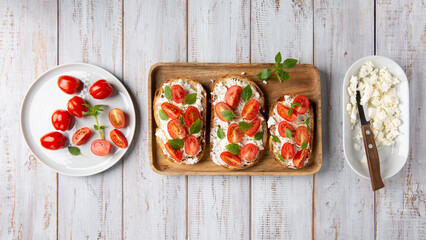 Sandwich with cottage cheese, tomatoes and basil on white wooden background. Traditional Italian bruschetta. Healthy savory feta and tomato toast. Top view. - obrazy, fototapety, plakaty