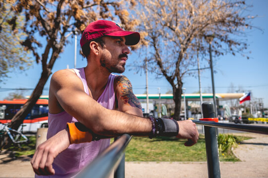 Concentrated, bearded, healthy and fit young latin man with colorful tattoo and cap resting a moment in parallel bars in a park in a sunny day
