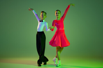 Beginner dancers. Portrait of beautiful little boy and girl dancing ballroom dance isolated over green background in neon light. Concept of art, beauty, grace, action, emotions.