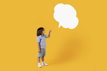 Interesting news. Cute little boy looking at blank speech bubble, pointing with finger, orange background, empty space