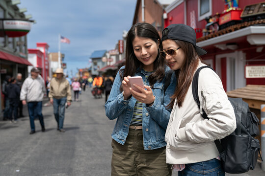 two asian Taiwanese girl friends looking at tourist map online on the phone while discussing travel plans together at Old Fishermanâs Wharf in California usa
