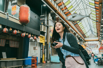 Plakat cheerful Chinese woman photographer enjoying view along the arcade while taking pictures in kuromon ichiba market in Osaka japan with fish hanging decoration at background