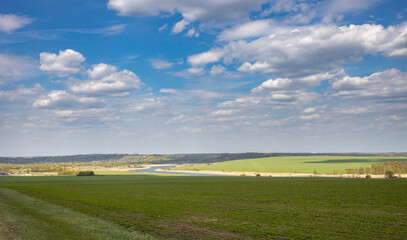 Sunlight through the clouds illuminates the young greenery of bushes and trees. Rural spring landscape with a river, clouds over the horizon.