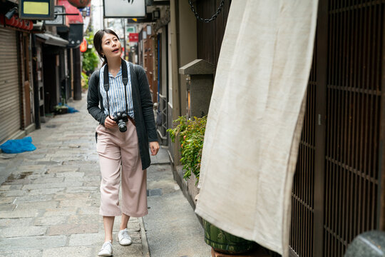 Asian Woman Tourist Holding Digital Camera Looking At White Door Curtain Outside A Japanese Restaurant While Walking In Narrow Paved Alley At Leisure In Nanba-eki Osaka Japan