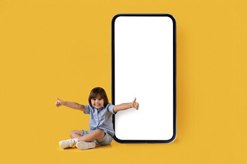 Content for kids. Excited little boy sitting near big smartphone with blank white screen and gesturing thumbs up