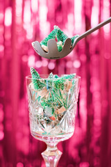 An antique glass goblet is filled with miniature Christmas trees against a backdrop of red glittery tinsel. The scoop adds a few more fancy trinkets to the glass. Humorous New Year concept.