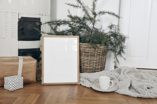 Festive Christmas indoor decor. Vertical wooden picture frame mockup on parquette floor. Pine, larch tree branches in basket, gift boxes. Cup of coffee. Old white tiled stove background.Empty template