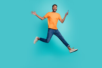 Fototapeta na wymiar Full length photo of excited positive guy wear orange t-shirt jumping high chatting modern gadget isolated teal color background