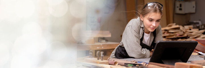 web banner Portrait of a female carpenter looking at designs on a tablet for making her furniture in a furniture factory. with many tools and wood with modern tools with copy space on left