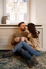 Fototapeta premium Romantic couple at home. An Attractive young woman and handsome man are enjoying spending time together while siting coddling with cups of tea in hands.