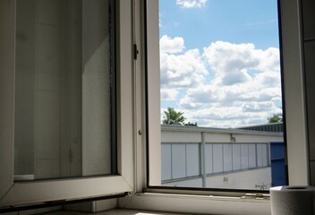 Offenes Fenster mit weißem Rahmen und Ausblick nach draußen zu weißem Fabrikgebäude vor blauem...