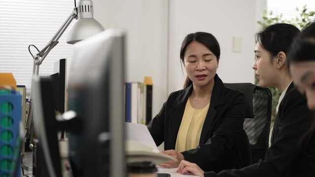selective focus of friendly female manager explaining new online project with paper and hand gesture to young female worker at office desk in the company