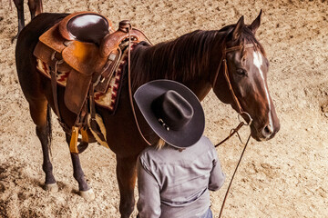 A Thoroughbred horse with a Western saddle standing with a person in a cowboy hat in an arena taken...