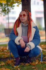 Young thoughtful woman with bright dreadlocks, sunglasses squatting near tree in park. Vivid female...