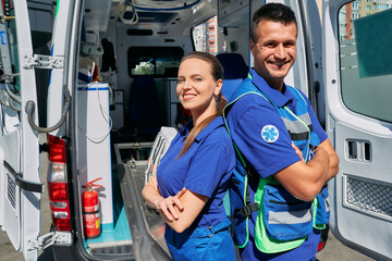 Confident paramedics team standing near their ambulance car with crossed arms. Personnel of emergency service